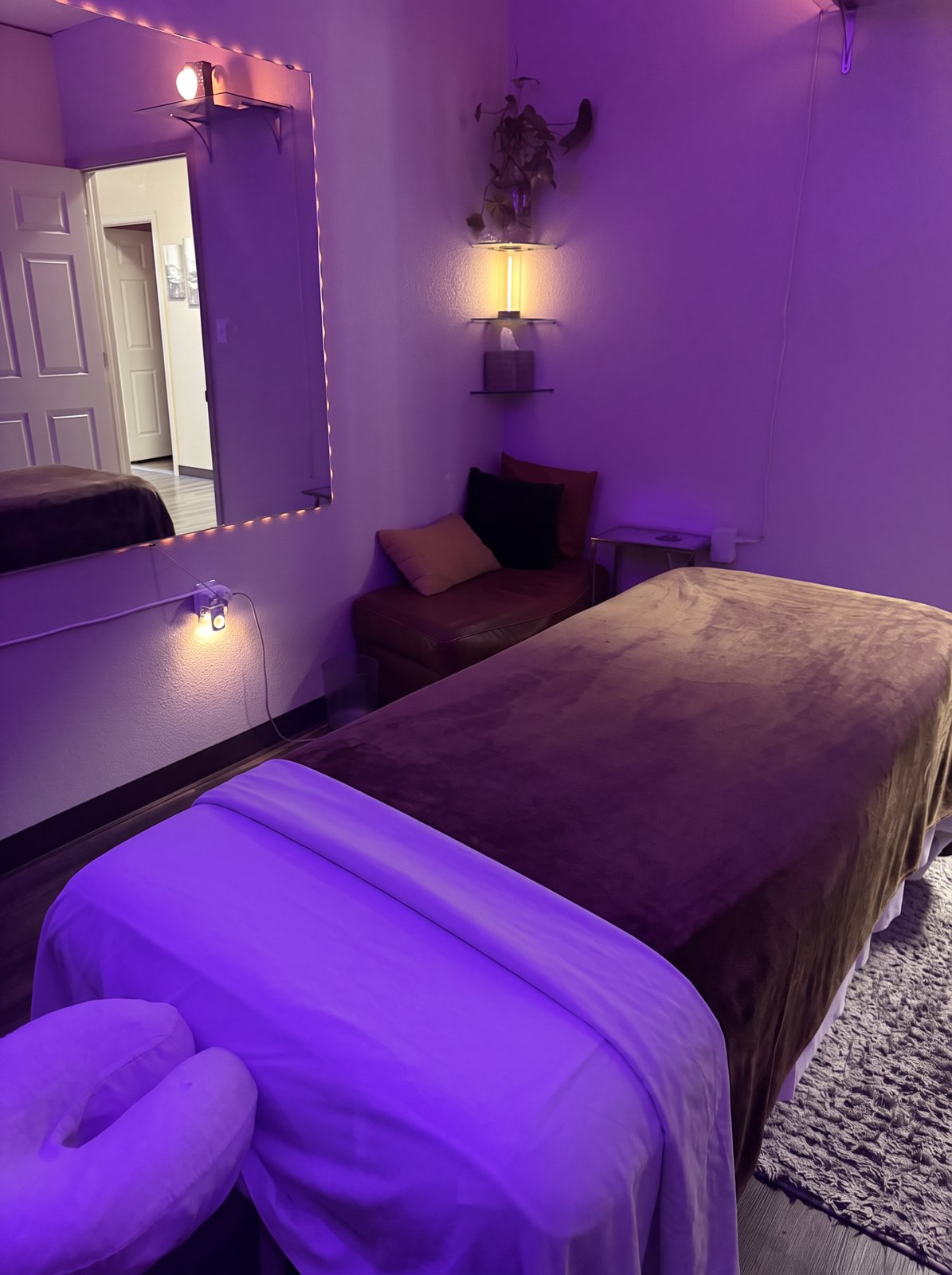 Jorgé’s massage table beside a light-framed mirror, chair with pillows, and wall shelves